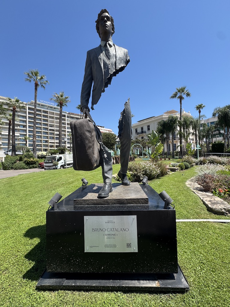 Fotoğraftaki bu etkileyici heykel, Fransız sanatçı Bruno Catalano’nun dünyaca ünlü "Les Voyageurs" (Gezginler) serisine ait "Simone" adlı eseri. Cannes’da, ünlü La Croisette üzerinde sergilenen bu bronz çalışma, ellerinde bir bavulla bilinmeze doğru adım atan, ancak gövdelerinin büyük bir kısmı tamamen "eksik" olan figürleri anlatıyor. Sanatçı, kendisi de bir göçmen olarak, köklerinden ayrılıp yeni bir yere seyahat eden insanların geride bıraktıkları boşluğu, hissettikleri eksikliği ve her yolculuğun insanın ruhundan bir parçayı koparıp götürüşünü bu çarpıcı boşluklarla simgeliyor. Havada asılı duruyormuş hissi veren heykel, fiziksel olarak parçalanmış görünse de elindeki bavul sayesinde üst ve alt gövdesini birbirine bağlayarak hem aidiyetsizliği hem de umuda doğru atılan o kararlı adımı gözler önüne seriyor.